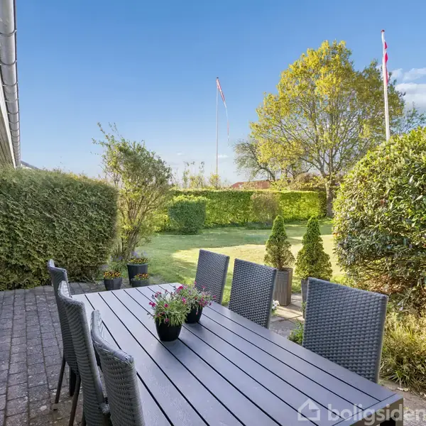 Et langt bord med kurvestole står på en flisebelagt terrasse. Terrassen er omgivet af en velplejet have med grønne buske og træer under en klar blå himmel. Text: boligsiden.