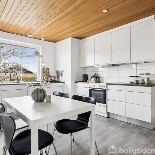 Et lyst køkken indeholder et hvidt bord med sorte stole placeret ved siden af en vindue. Hvide skabe og en ovn omkranser rummet, mens et træloft tilføjer varme.