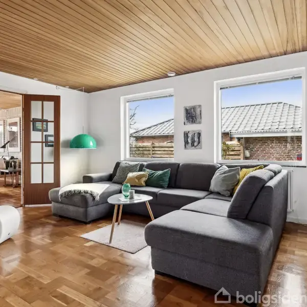 En grå hjørnesofa står i en lys stue med parketgulv. Ved siden af sofaen er et rundt bord med en lampe. To store vinduer viser en udendørs mur og tag.