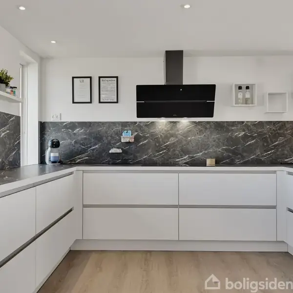 Hvidt moderne køkken med mørk marmorbordplade og backsplash, indbyggede skabe, og sort emhætte. Lysindfald fra vindue. Hylder med planter og indrammede billeder på væggen. Ingen tekst er synlig.