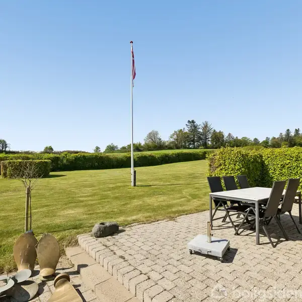 Et bord med fire stole står på en terrasse med fliser. En flagstang med et dansk flag står på en velplejet græsplæne omgivet af en hæk og træer i baggrunden.