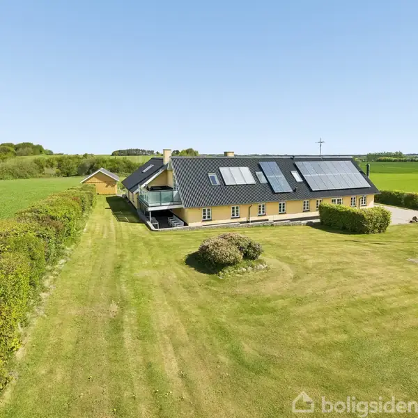 Hus med solpaneler på taget; omgivet af velplejet græsplæne og buske; dansk flag i forgrunden; landlig baggrund med marker og hække; blå himmel uden tekst.