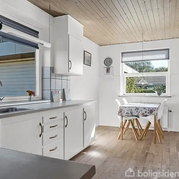 Et køkken med hvide skabe og trækøkkenbord, hvor en vask er placeret. Ved siden af er et lille spisebord med to stole. Store vinduer slipper dagslys ind.