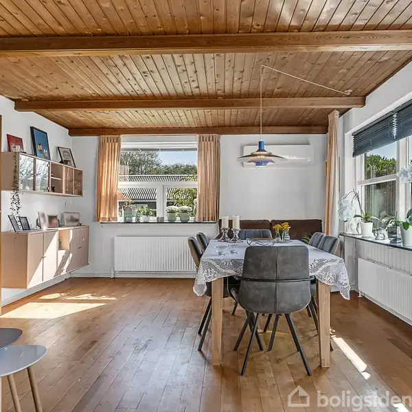 Spisebord omgivet af seks stole står i et lyst rum med trælofter. På bordet er en dug og en lysestage. Væggene har store vinduer med planter og hvide radiatorer.