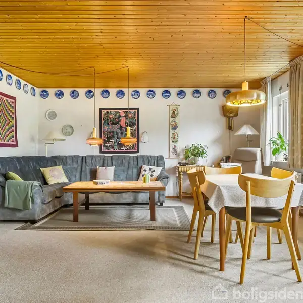 Stue med grå sofa, træbord, spisebord med fire stole, dekoreret med blå tallerkner på væggen, grønne planter, lamper over bordet, lys træloft og store vinduer med gardiner.