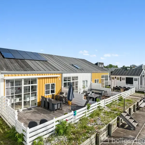 Et gult træhus med solpaneler på taget står ved siden af en terrasse med møbler. Huset er omkranset af en lav hvid hegn, og omgivet af andre lignende boliger.