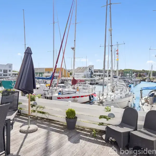 En terrasse med sorte havemøbler står stille, omgivet af en marina med sejlbåde i vandet. Bygninger i forskellige farver ses i baggrunden under en blå himmel.