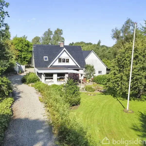 Et hvidt hus med sort tag står blandt grønne træer og have. En grusvej fører op til huset, omgivet af velplejede buske og en flagstang på plænen.