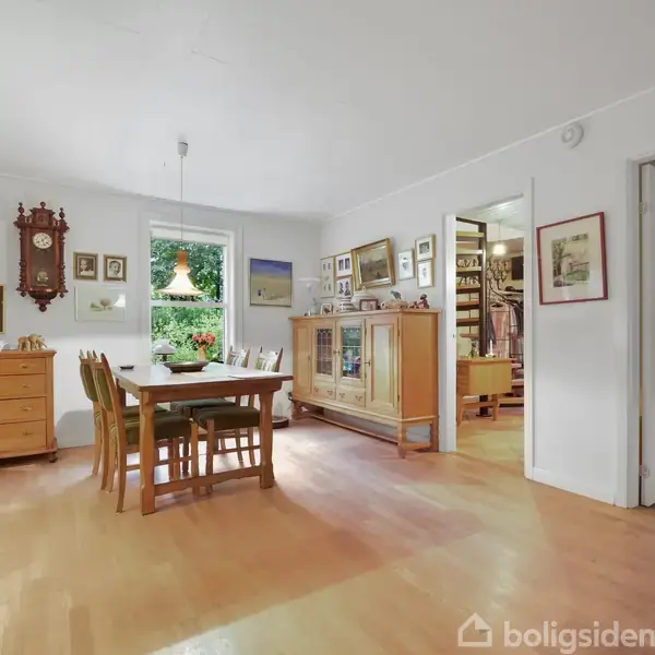 Et spisebord med stole står i midten af et lyst rum. Rummet har en trækommode, vægure og flere billeder på væggene. En dør åbner op til et andet rum.