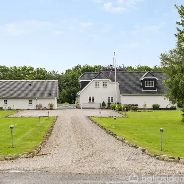 Et hvidt rækværkshus med sort tag ligger i baggrunden, flankeret af to mindre bygninger. En grusvej fører op til huset, omgivet af velplejet græsplæne og buske.