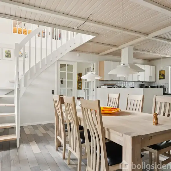 Et spisebord med syv stole står i et lyst, moderne køkken-alrum med trægulve. En hvid trappe fører op til en overetage, og rummet er indrettet med minimalistiske møbler.
