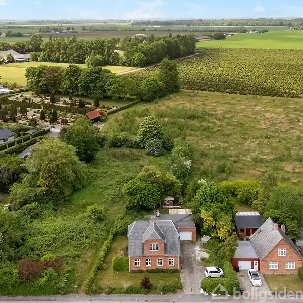 To huse med røde tage står tæt sammen omgivet af frodige træer og buske. En mark strækker sig i baggrunden, mens flere huse ligger på den anden side.
