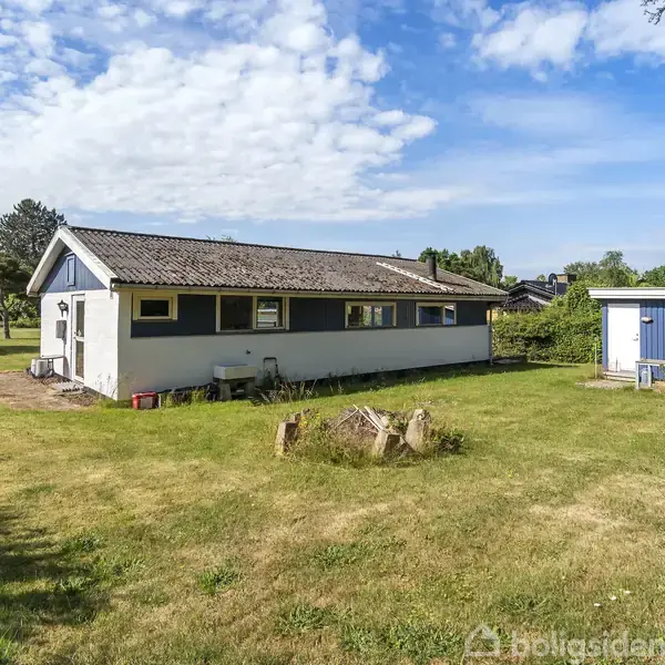 En enkeltstående, hvid bygning ligger stille på en grøn græsplæne med en mindre, blå udhus ved siden af. Det er omgivet af buske og træer under en blå himmel.