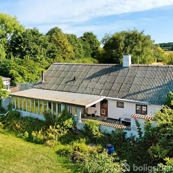 Huset, med skråt tag og hvide vægge, ligger omgivet af frodig grøn have og træer. Det har en lang veranda og er placeret i et landligt område ved siden af en mark.