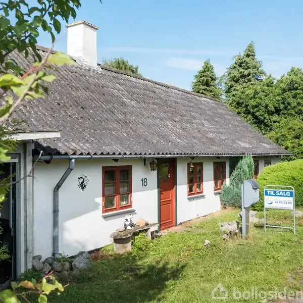 Et hvidt hus med stråtag står langs en vej omgivet af grønt løv. Et skilt ved vejen siger "Til salg" med et telefonnummer 63 15 15 15.