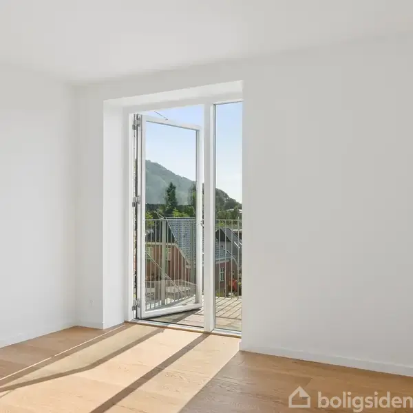 Et lyst rum med trægulv og en åben dør fører ud til en balkon. Udenfor ses en bjergudsigt og blå himmel. Ingen tekst i billedet.