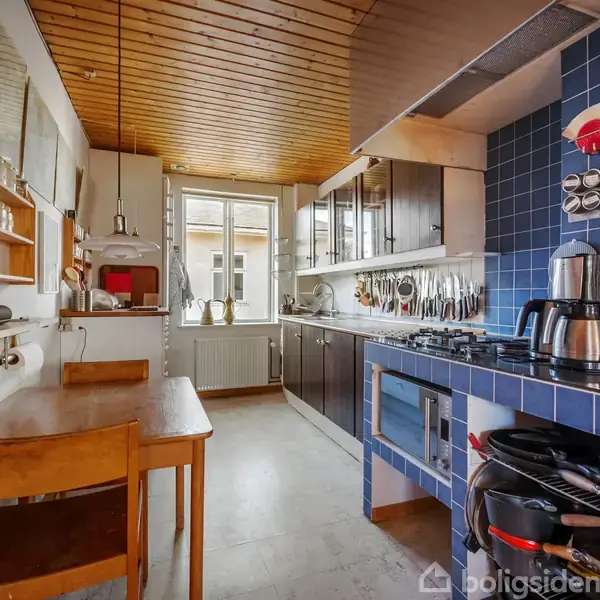Et køkken med træloft og blå fliser, hvor der ses en komfur og mikroovn. Bord og stole står ved siden af en hylde med krydderiglas. Vindue for enden giver lysindfald.