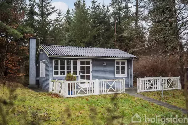 Et lille, blåmalet sommerhus med hvidt stakit står i en skovklædt have. Taget er af bølgeblik, og huset er omgivet af græsplæne og træer.