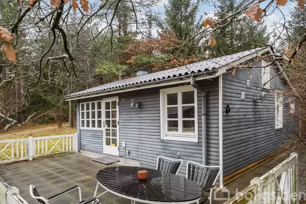 Træhus med grå facade står stille i skovklædt område. Terrasse foran huset har rundt bord med stole. Omgivet af hegn og efterårsløv.