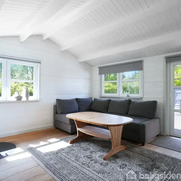 En stue med en hjørnesofa og et træbord står på et tæppe nær et vindue. Pejsen til venstre opvarmer rummet, som har hvide trævægge og lyse trægulve.