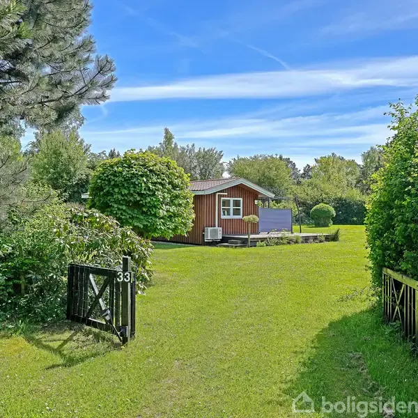 Et træhus står i en solrig have omkranset af træer og buske, med en åben grøn låge markeret med tallet "33" forrest i billedet.