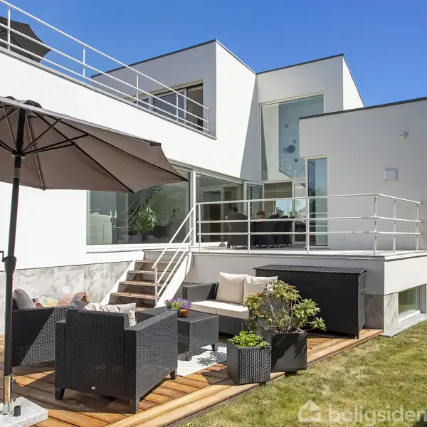 Et moderne hvidt hus med store vinduer har en terrasse med sorte møbler og planter. En stor parasol skygger over en del af terrassen. Græsplæne omgiver området.