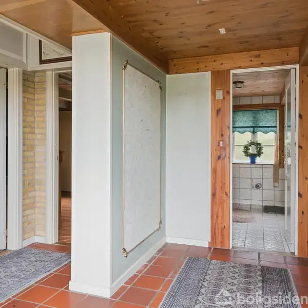 Korridor med flisegulv og tæpper, åbner ud til flere døre og badeværelse i enden. Væggene har lyse farver og trædetaljer, mens vinduet har en grøn rullegardin og dekorativ plante.