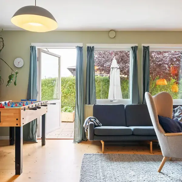 Fodboldbord står i et lyst rum. En reol med bøger og planter i venstre side. Sofa og lænestol ved vinduer med udsigt til have.