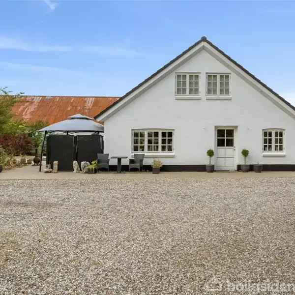 Et hvidt hus med trempel tag står foran en grusbelagt gårdsplads. Ved siden af huset er der havemøbler og grønne planter. Omkring er der træer og buske.