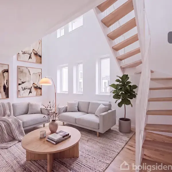 Stue med to grå sofaer og rundt træbord, dekoreret med bøger og en vase. En stueplante ved siden af trapperne. Kunstværker hænger på væggen. Tekst: "Visualisering".