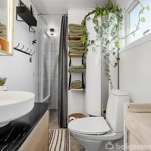 Stue med en stor, grå hjørnesofa, en brændeovn i hjørnet, og runde sofaborde. Vinduet viser træer udenfor. vægdekorationer og en hængende lampe tilføjer stil.