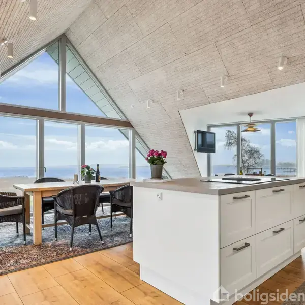 Et moderne køkken med en hvid ø kigger ud mod havudsigt gennem store glasvinduer. Et spisebord med fire stole står ved vinduerne, omgivet af træ- og tæppebelagt gulv.