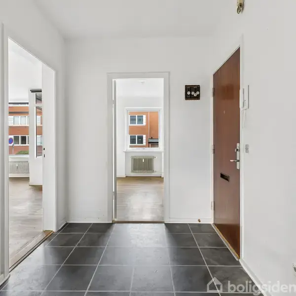 Entré med mørke fliser på gulvet, en brun dør til højre, åbning til to lyse værelser med store vinduer i baggrunden, som giver udsigt til en rød murstensbygning.