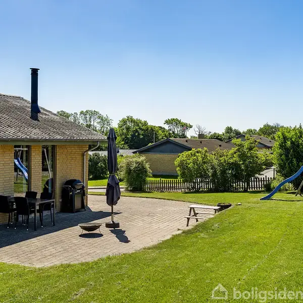 Hus med terrasse indeholder havemøbler og parasol. I haven står en legeplads med rutsjebane. Solrig dag i et villakvarter.