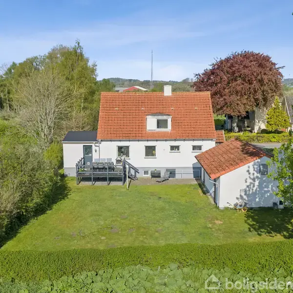 Et hvidt hus med rødt tag står i en grøn have. En lille tilbygning er ved siden af. Omkring er der træer og buske i et landligt område.