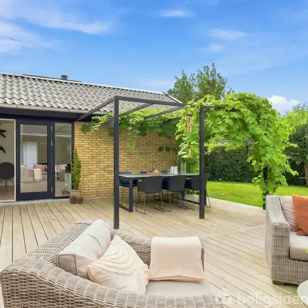 Terrassen har flettede møbler med puder, trædæk og spisebord under en pergola. Murstenshuset har glasdøre, omgivet af grønne planter i en velplejet have med blå himmel.