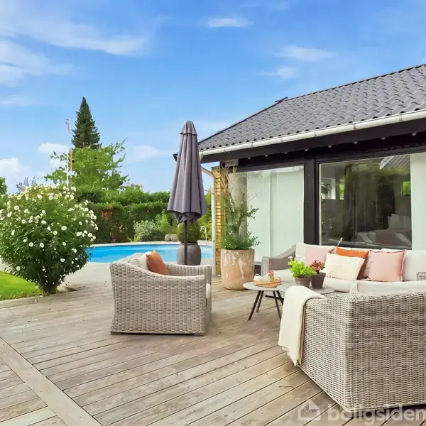 Terrasseområde med møbler og parasol ved siden af et hus med store vinduer. I baggrunden ses en swimmingpool og grøn have med blomstrende buske under en klar blå himmel.