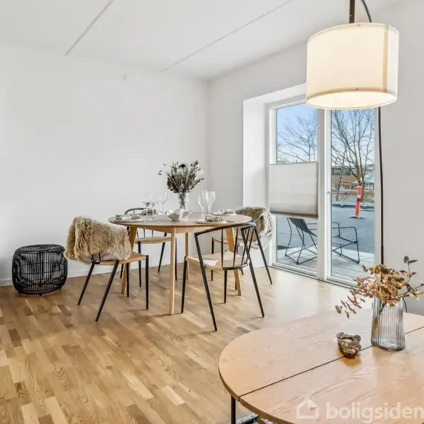 Et spisebord står dækket med glas og servietter i en lys stue med trægulv. Store vinduer lader naturligt lys strømme ind. En hængelampe lyser rummet.