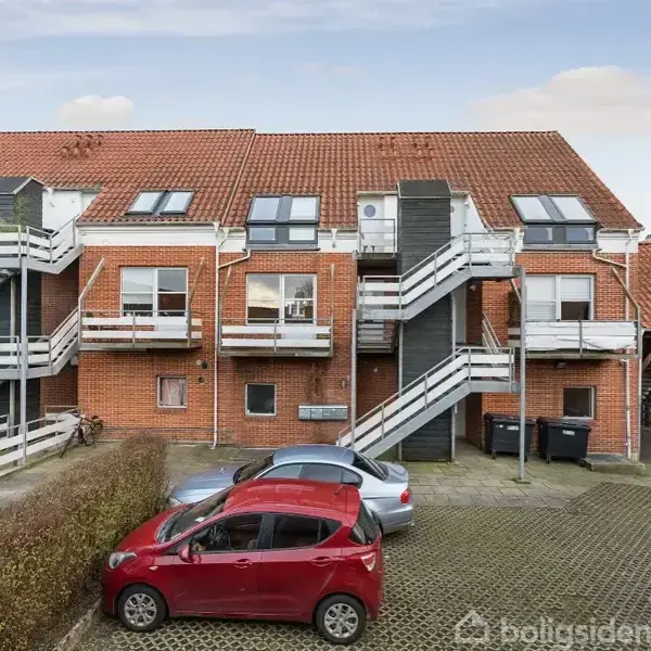 Røde murstenshuse med flere eksterne trapper fører til andet niveau og loft. To biler, en rød og en sølv, er parkeret foran husene på en græs- og flisebelagt parkeringsplads.