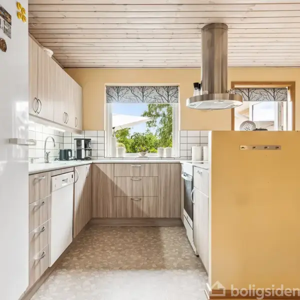 Køkken med hvide apparater og træpaneler. Emhætte over kogeplade, vinduer med udsigt til grønt udenfor. Bord og stole i spiseområde til højre. Lysindfald fra vinduer.