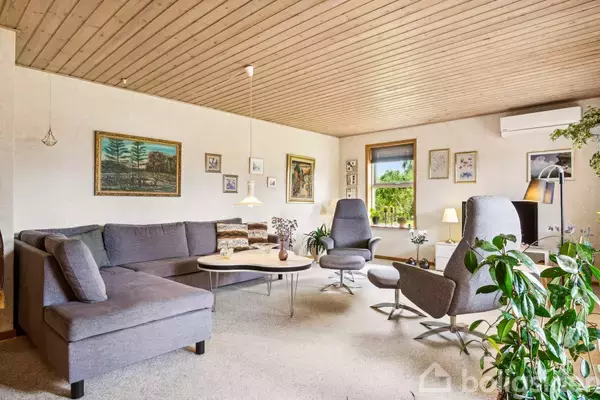 Grå hjørnesofa står i stue med flere billeder på væggene. To grå lænestole ved kaffebord. Store vinduer viser grøn udsigt. Indendørs planter tilføjer naturligt præg.