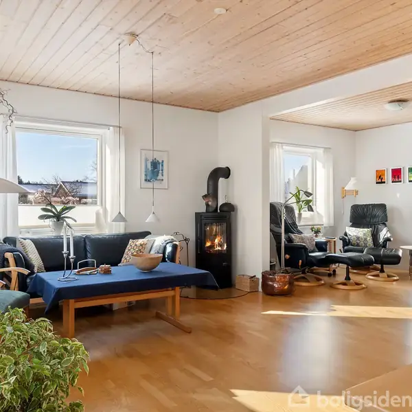 Stue med sofa og kamin i centrum, træloft og lyse vægge. Et piano og lænestole er placeret ved siden af store vinduer. Indretning med farverige billeder og planter.