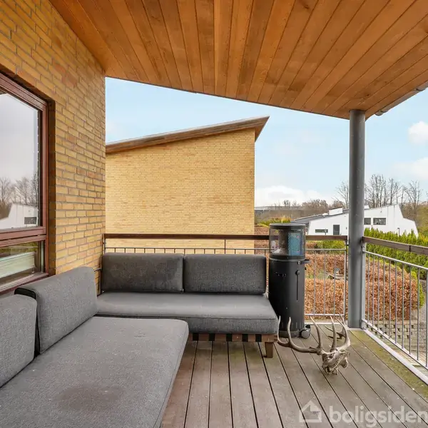 Grå sofaer står på en overdækket altan med træterrasse. En varmelampe er til stede. Ombygninger og træer kan ses i det omkringliggende boligområde.