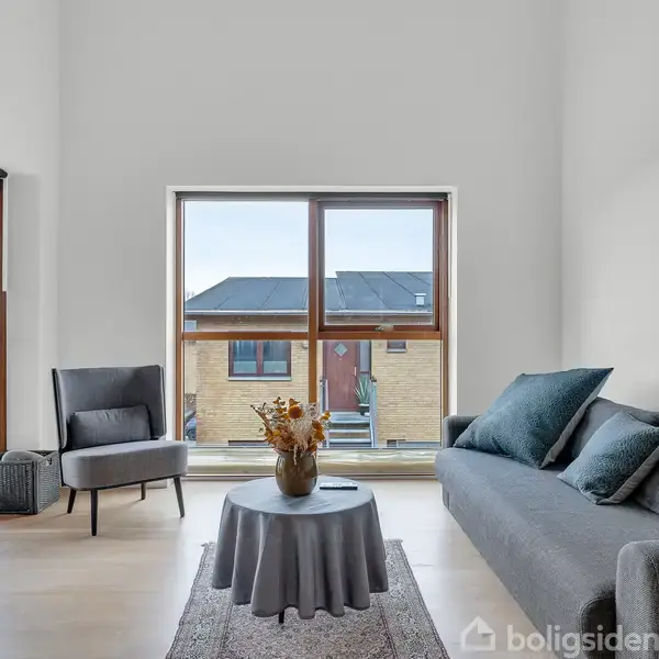Et lyst rum med en grå sofa og stol omkring et lavt bord med blomster. Store vinduer giver udsigt til nabohusene. En dør til venstre fører ud.