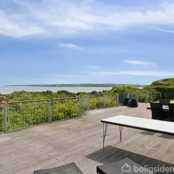 Stor terrasse med bord og stole står på solrig træoverflade. Omgivet af grønne buske og træer, beliggende ved kysten med udsigt over havet og en blå himmel.