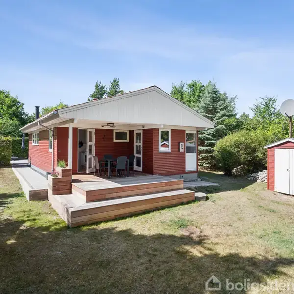 Et rødt sommerhus med en overdækket terrasse har stole og et bord. Det ligger i en grøn have med træer og en lille rød udhusbygning med parabol.