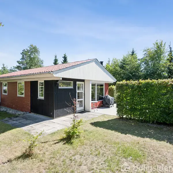 Et lille, rødt murstenshus med trædetaljer står i en have med græs og buske. Sollys skinner på terrassen, omgivet af frodig vegetation.