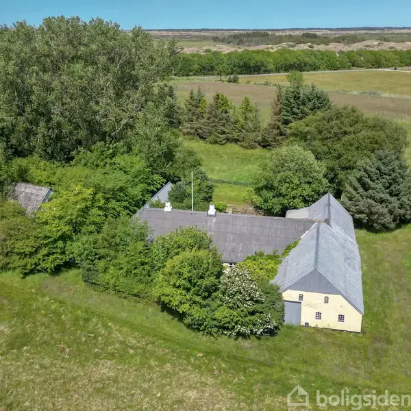 En gul bygning med gråt tag omgivet af tæt, frodig vegetation står i et åbent landbrugsområde med træer i baggrunden og marker, der strækker sig ud til horisonten.