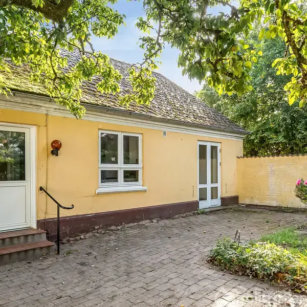 Et gult hus med skråt tag står i sollys. En glasdør og et vindue er synlige. En stenbelagt terrasse foran, omgivet af grønne træer og planter.