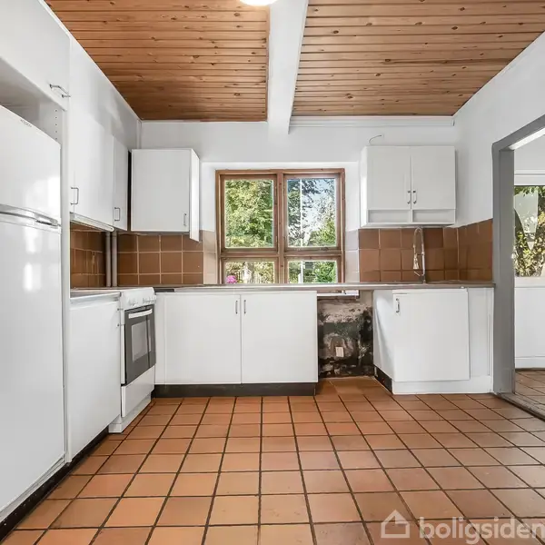 Køkken med hvide skabe og apparater, herunder komfur og køleskab. Rummet har brune fliser på gulvet og vægge samt et træloft. Et vindue viser udsigt til grøn vegetation.